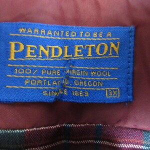 Puede incluir: Una etiqueta azul de Pendleton con letras doradas, cosida sobre una tela burdeos. La etiqueta dice "WARRANTED TO BE A PENDLETON 100% PURE VIRGIN WOOL PORTLAND, OREGON SINCE 1863 3X". Se aprecia un patr&oacute;n de cuadros en el fondo.