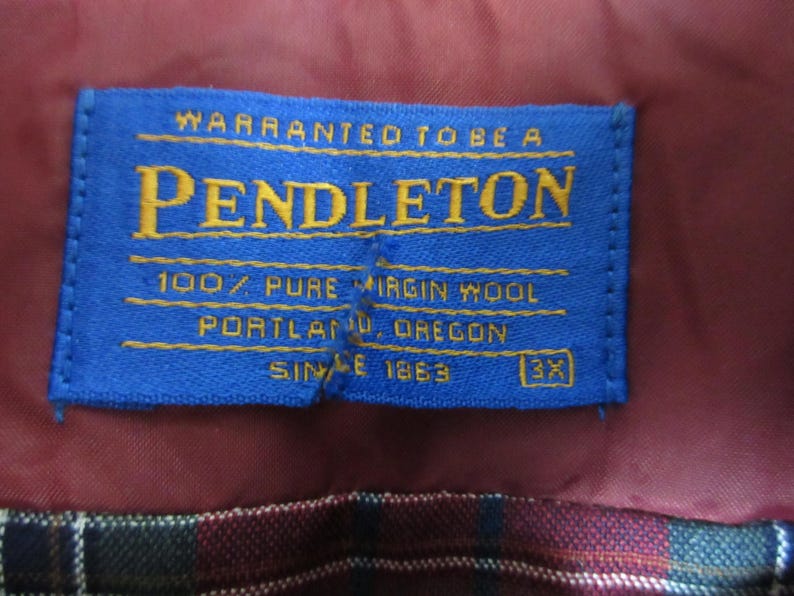 Puede incluir: Una etiqueta azul de Pendleton con letras doradas, cosida sobre una tela burdeos. La etiqueta dice "WARRANTED TO BE A PENDLETON 100% PURE VIRGIN WOOL PORTLAND, OREGON SINCE 1863 3X". Se aprecia un patr&oacute;n de cuadros en el fondo.