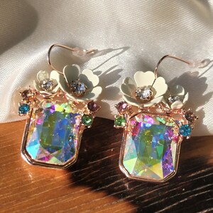 May include: A pair of iridescent gemstone earrings with a floral design. The earrings are made of gold-toned metal and feature a large, rectangular gemstone in the center. The gemstones are surrounded by smaller, colorful gemstones and white flowers.