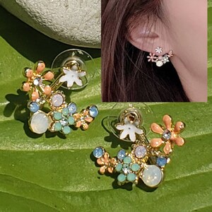 Puede incluir: Un par de pendientes de tono dorado con un diseño floral. Los pendientes presentan un grupo de flores de color rosa pastel, azul y blanco con pedrería transparente. Los pendientes están diseñados para llevarse en la parte superior de la oreja.