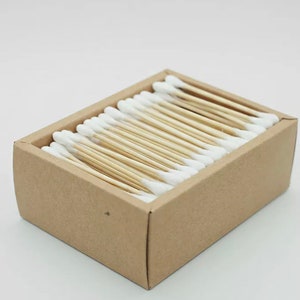 May include: A rectangular cardboard box filled with cotton swabs. The swabs have white cotton tips and light brown wooden stems. The box is a light brown color and is sitting on a white surface.
