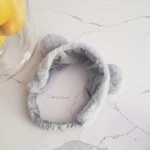 May include: A gray fuzzy headband with two fluffy ear-like accents. The headband is made of soft, plush material.