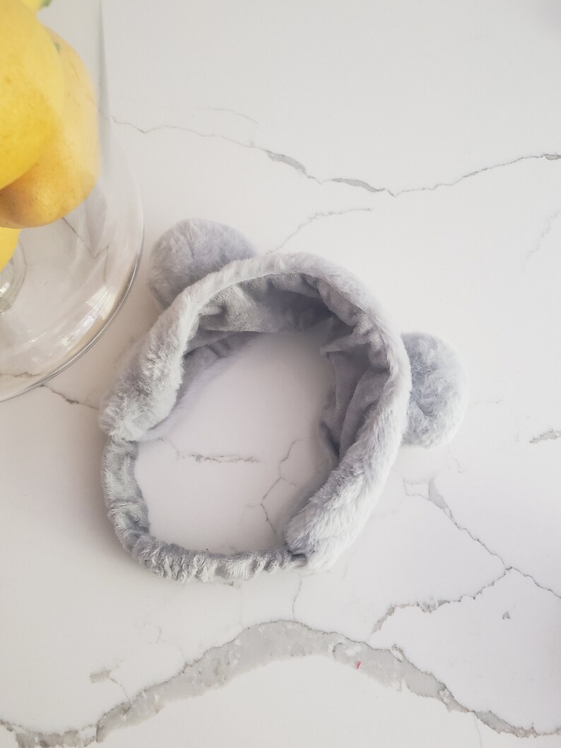 May include: A gray fuzzy headband with two fluffy ear-like accents. The headband is made of soft, plush material.