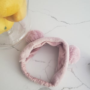 May include: A pink fuzzy headband with two fluffy pom-poms on the sides. The headband is made of soft, plush material.