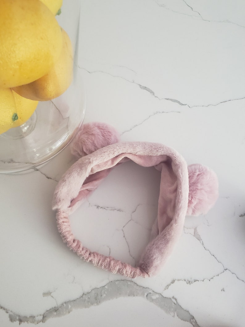 May include: A pink fuzzy headband with two fluffy pom-poms on the sides. The headband is made of soft, plush material.