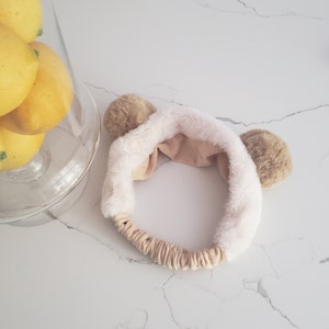 May include: A white and brown fuzzy headband with two brown bear ears. The headband is made of soft, plush fabric.