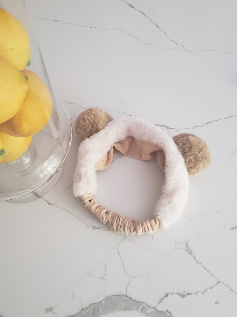 May include: A white and brown fuzzy headband with two brown bear ears. The headband is made of soft, plush fabric.