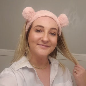 May include: A woman wearing a white button-down shirt with a gold chain necklace. She is wearing a pink fuzzy headband with bear ears. Her hair is blonde and she is holding a strand of her hair in her right hand.