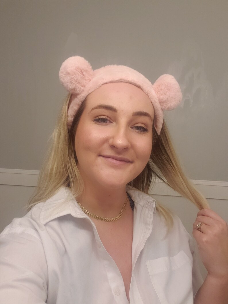 May include: A woman wearing a white button-down shirt with a gold chain necklace. She is wearing a pink fuzzy headband with bear ears. Her hair is blonde and she is holding a strand of her hair in her right hand.