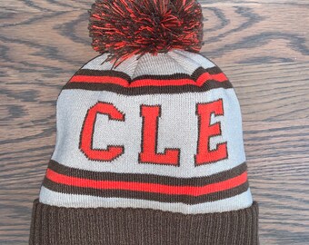 May include: A brown and gray knit beanie with a brown pom pom on top. The beanie has a white stripe with the word "CLE" in orange letters.