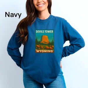 May include: Navy blue long-sleeve shirt featuring a Devils Tower, Wyoming graphic. The graphic includes the text "Devils Tower" and "Wyoming" in a rectangular frame. The shirt is made of a soft, comfortable material.