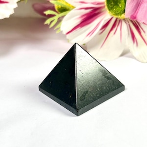 May include: A small, black pyramid made of a solid material. The pyramid has a square base and four triangular sides that meet at a point. The pyramid is set against a white background with pink and white flowers.