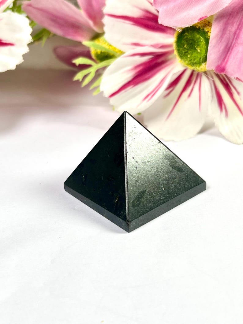May include: A small, black pyramid made of a solid material. The pyramid has a square base and four triangular sides that meet at a point. The pyramid is set against a white background with pink and white flowers.