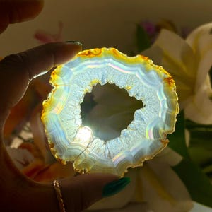 May include: A translucent, circular agate geode slice with a central opening. The geode features bands of yellow, orange, and white, with iridescent qualities. The geode is held up, allowing light to pass through, highlighting its natural patterns and colours.