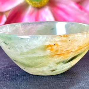 May include: A small, translucent bowl with a rounded shape. The bowl features a blend of green, yellow, and white colors, with a polished, glossy finish. The bowl is set against a blurred background of pink and green.