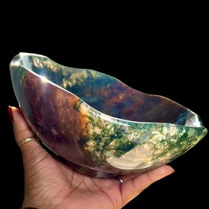 Puede incluir: Un cuenco decorativo con un patrón natural único. El cuenco presenta una mezcla de tonos verdes, marrones y azules, creando un efecto veteado. El borde tiene un diseño ondulado. El cuenco se sostiene en una mano sobre un fondo negro.