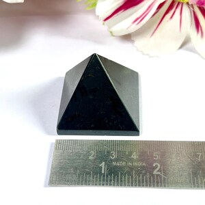 May include: A small, black pyramid-shaped stone, approximately 2 inches wide at the base, sits on a white surface. The pyramid is made of a dark, possibly polished material. A metal ruler is placed next to the pyramid for scale. Decorative flowers are in the background.