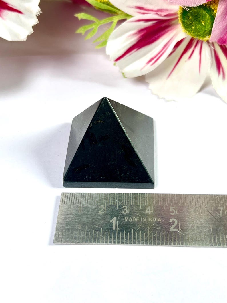 May include: A small, black pyramid-shaped stone, approximately 2 inches wide at the base, sits on a white surface. The pyramid is made of a dark, possibly polished material. A metal ruler is placed next to the pyramid for scale. Decorative flowers are in the background.