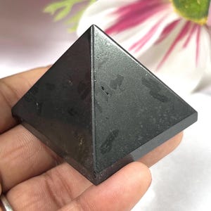May include: A dark gray pyramid-shaped stone, held in a hand. The pyramid has a flat base and four triangular sides that meet at a point. The stone has a slightly textured surface.