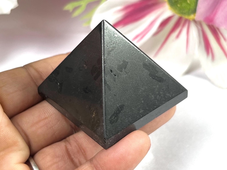 May include: A dark gray pyramid-shaped stone, held in a hand. The pyramid has a flat base and four triangular sides that meet at a point. The stone has a slightly textured surface.