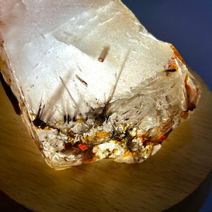 May include: A translucent, rough-cut crystal with internal inclusions and a warm, glowing light. The crystal features streaks of black and reddish-orange hues, set against a clear, textured background. The crystal is displayed on a wooden base.