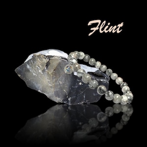 以下が含まれることがあります： グレーと白の模様のビーズブレスレットが、黒い背景に暗い灰色の岩の上に置かれています。「Flint」という単語が、岩とブレスレットの上に筆記体で書かれています。