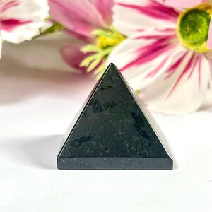 May include: A small, black pyramid-shaped stone with a flat base. The pyramid has a smooth, polished surface and is set against a backdrop of pink and white flowers. The pyramid is a decorative item.