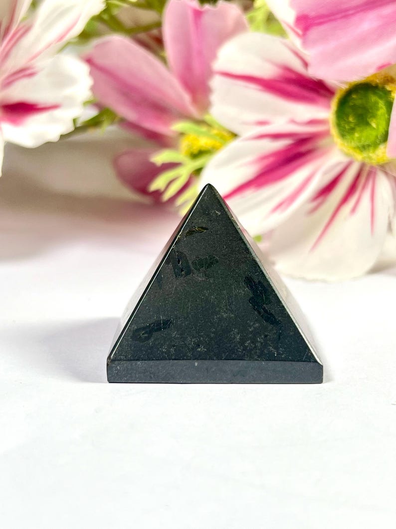 May include: A small, black pyramid-shaped stone with a flat base. The pyramid has a smooth, polished surface and is set against a backdrop of pink and white flowers. The pyramid is a decorative item.