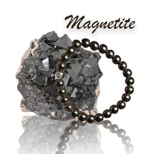 Puede incluir: Una pulsera de magnetita gris oscuro con cuentas esféricas pulidas se muestra junto a una piedra de magnetita gris oscuro y rugosa. La palabra "Magnetite" está escrita en cursiva encima de la pulsera y la piedra.