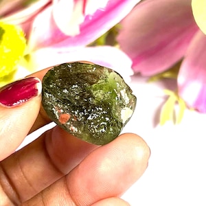 May include: A rough, green gemstone with a textured surface. The stone has small, reddish-brown inclusions and is held against a backdrop of pink and yellow flowers. The gemstone is approximately 1 inch in length.