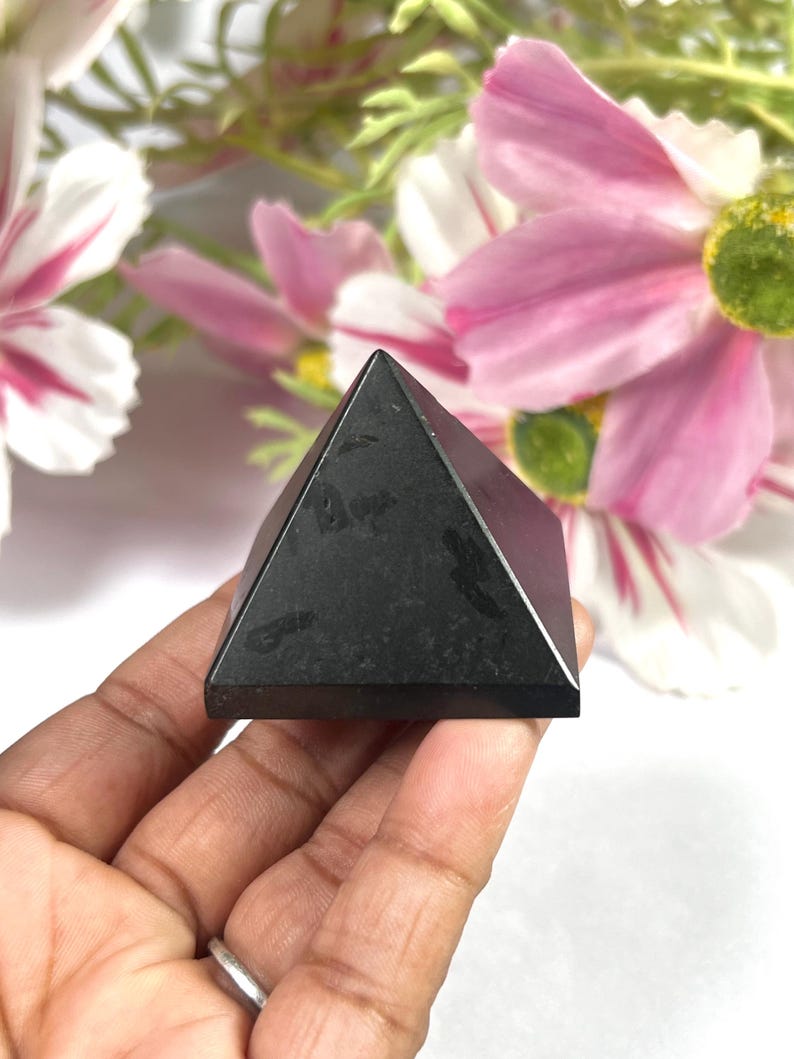 May include: A small, black pyramid-shaped object with a smooth surface. The pyramid is held in a hand, with a blurred floral background of pink and white flowers. The pyramid's shape is a classic geometric form.