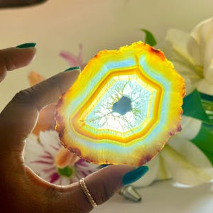 May include: A translucent, colorful agate slice with a central hole. The stone features bands of yellow, orange, and light blue hues. The agate is held up by a hand with teal nail polish, with a gold ring on the finger.