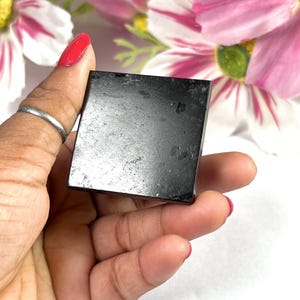 May include: A small, square, black stone held in a hand with red nail polish. The stone has a slightly textured surface. Pink and white flowers are in the background.