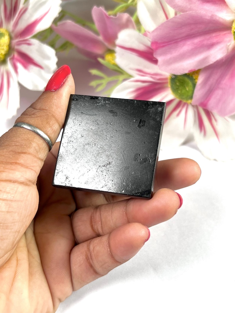 May include: A small, square, black stone held in a hand with red nail polish. The stone has a slightly textured surface. Pink and white flowers are in the background.