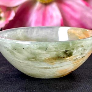 May include: A small, translucent bowl with a rounded shape. The bowl features a blend of green, white, and tan hues, creating a marbled effect. The bowl is likely made of stone or a similar material, and is suitable for holding small items.