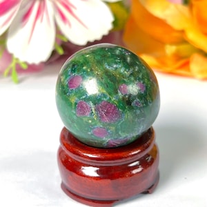 Peut inclure: Une sphère verte polie avec des inclusions rouges, posée sur un petit socle rond en bois brun rougeâtre. La surface de la sphère est lisse et réfléchissante, avec des inclusions rouges dispersées. En arrière-plan, des fleurs floues roses et oranges.