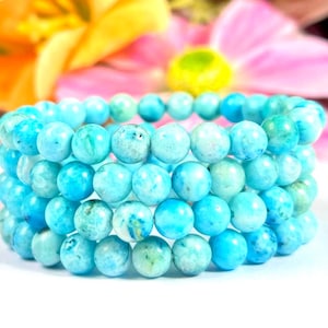 May include: Three stacked bracelets made of round, turquoise-colored beads. The beads have a marbled appearance with shades of blue and green. The bracelets are displayed against a soft, blurred floral background.