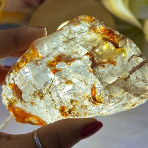 May include: A translucent, irregularly shaped gemstone with orange and brown inclusions. The stone is held in a hand with a silver ring, and is illuminated from behind, creating a glowing effect. The background is blurred, with hints of flowers.