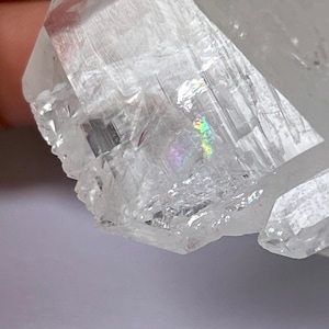 Puede incluir: Un cristal de cuarzo transparente con una iridiscencia arcoíris. El cristal está facetado y tiene una punta afilada.