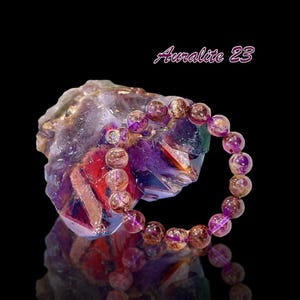 May include: An Auralite 23 bracelet made with purple, gold, and brown beads. The bracelet is resting on a large amethyst crystal.