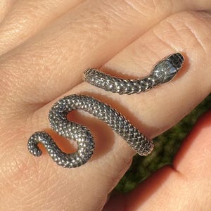 Può includere: Un anello a forma di serpente color argento con una superficie strutturata. La testa del serpente presenta una piccola pietra trasparente. L'anello è avvolto attorno a un dito, con il corpo del serpente che forma un design curvo.