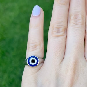 May include: A silver ring featuring a blue and white evil eye charm. The ring is worn on a finger with light purple nail polish.