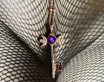May include: A bronze-colored pendant necklace featuring a key design. The key has a purple gemstone center and crescent moon accents. The necklace hangs on a delicate chain with small bead details.