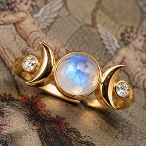May include: A gold ring featuring a central round iridescent moonstone cabochon flanked by two gold crescent moon shapes and two small clear round faceted stones set into the band on either side of the crescent moon accents.