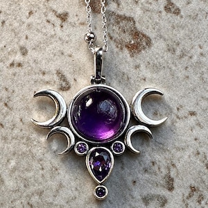 May include: A silver pendant necklace with a large, round, deep purple gemstone at its centre. The pendant features a triple moon design with crescent moons and smaller purple gemstones. The necklace chain is delicate.