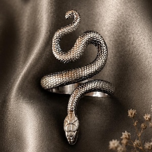 May include: A silver-colored snake ring with detailed scales and a coiled design. The snake's head is at the bottom, and the body curves upwards. The ring is set against a dark, silky background, with small white flowers.