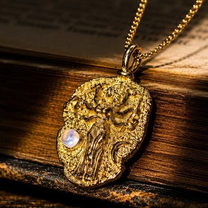 Puede incluir: Un collar con colgante en tono dorado que presenta una forma ovalada irregular y texturizada con una figura en relieve y un pequeño acento de piedra lunar redonda e iridiscente. El colgante cuelga de una delicada cadena dorada.