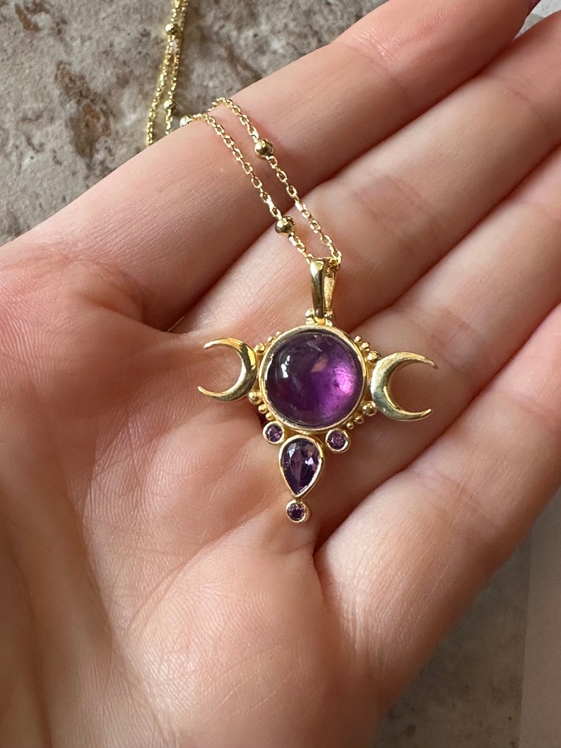 May include: Gold necklace with a pendant featuring a large, round, purple gemstone at the centre, flanked by crescent moons. Smaller purple gemstones are set below. The necklace has a delicate chain.