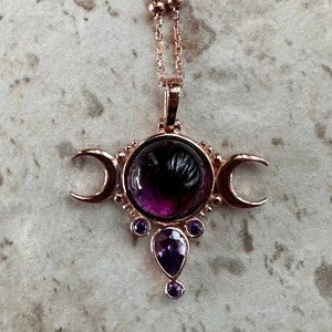 May include: Rose gold necklace with a central, large, round, deep purple gemstone, flanked by crescent moons and smaller purple gems. The pendant is suspended from a delicate chain with small, round beads.