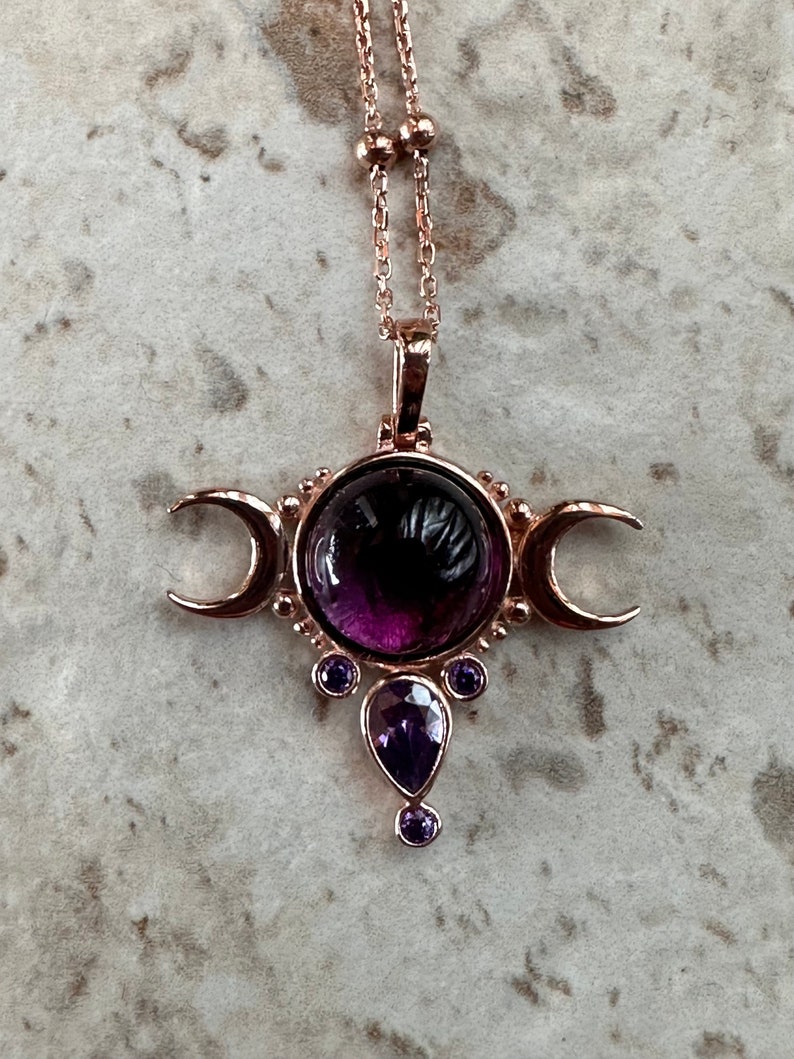 May include: Rose gold necklace with a central, large, round, deep purple gemstone, flanked by crescent moons and smaller purple gems. The pendant is suspended from a delicate chain with small, round beads.